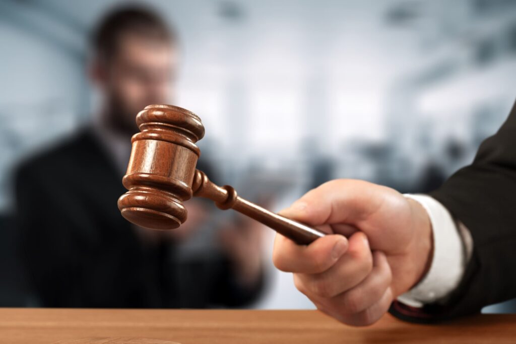 A judge’s gavel held forward by a person in a suit, with a courtroom blurred in the background.
